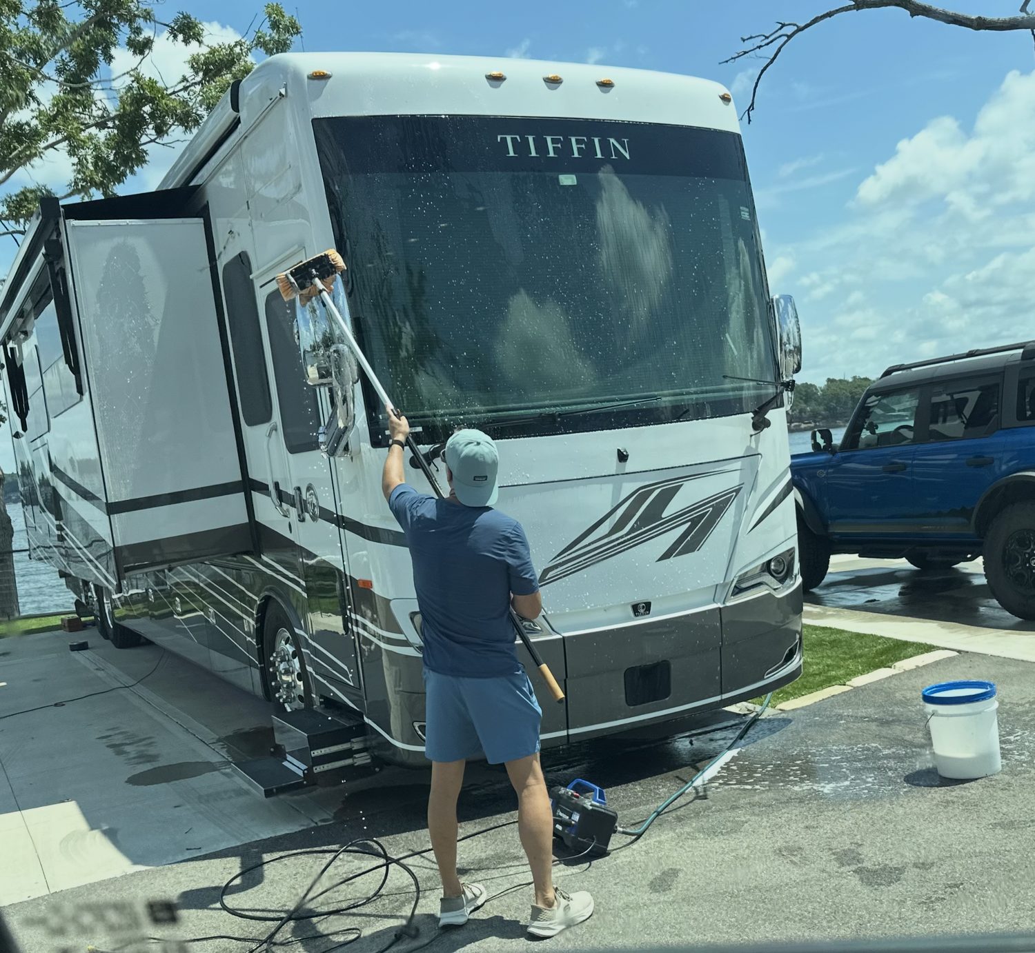 An owner scrubbing a Tiffin luxury coach windshield with a pole brush in the hot sun, tangled hoses and a portable pressure washer on the ground